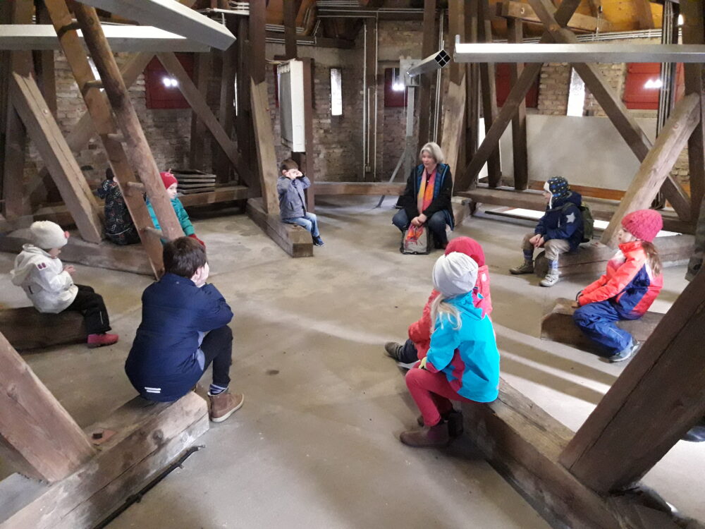 Eine Gruppe von Kindern sitzt auf Holzbänken in einem Raum mit sichtbaren Holzstützen. Einige Kinder tragen Mützen und Jacken in verschiedenen Farben.