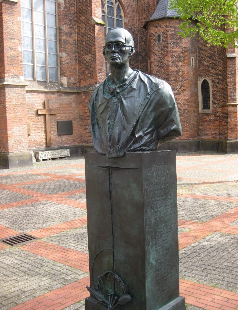 Bronzestatue von Karl Leisner, stehend im Vordergrund. Im Hintergrund die Stiftskirche mit roten Ziegelsteinen und einem hohen Kirchturm.