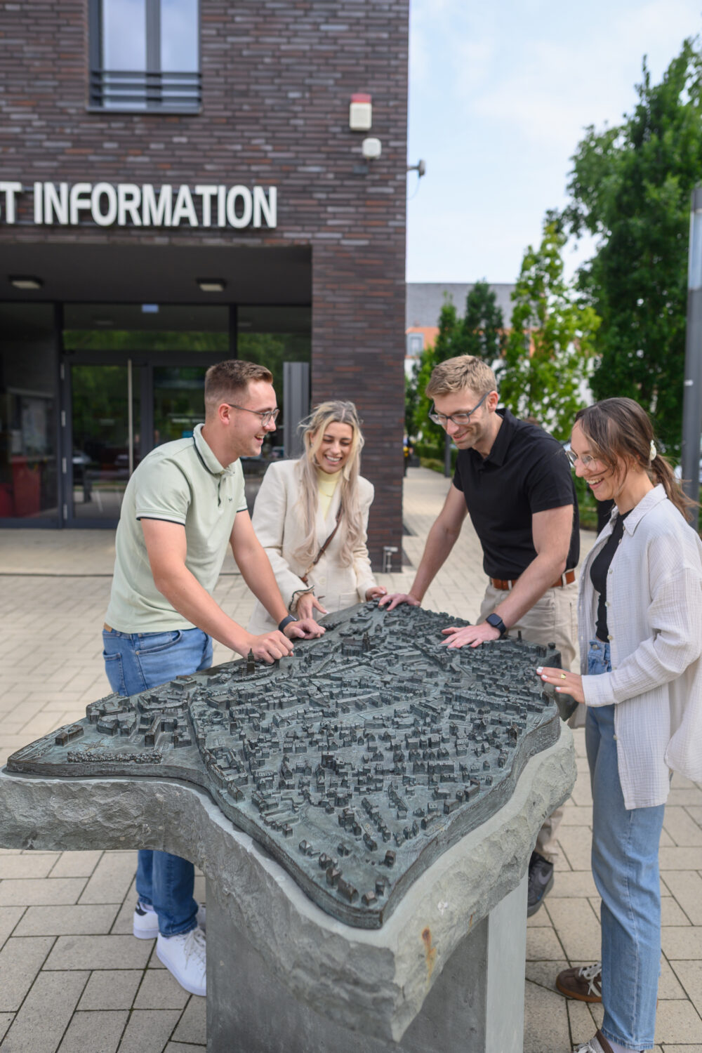 Vier Personen stehen um ein Stadtmodell aus grauem Stein, das eine detaillierte Karte zeigt. Im Hintergrund ist ein Gebäude mit dem Schriftzug 'TOURIST INFORMATION' zu sehen.