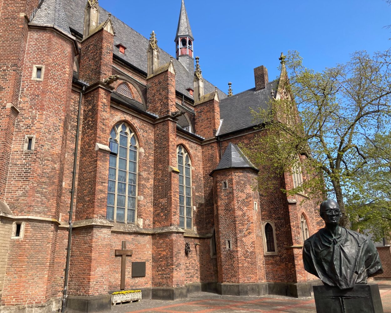 Die Stiftskirche in Kleve, aus rotem Backstein mit mehreren hohen Fenstern und einem spitzen Turm. Im Vordergrund steht eine Statue.