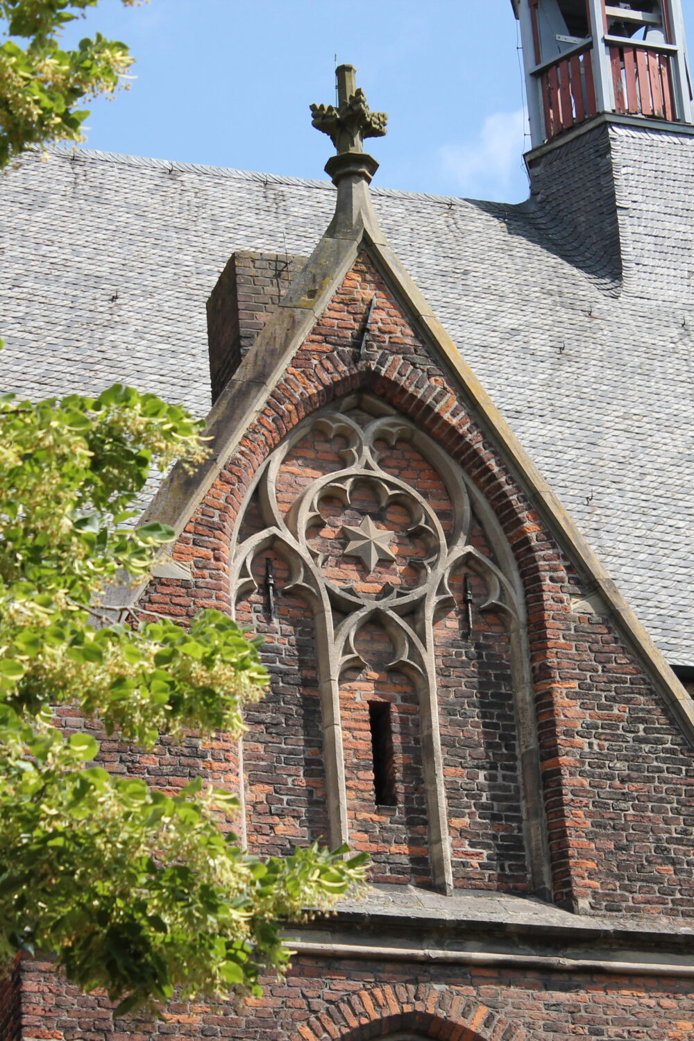 Detailansicht der Stiftskirche mit einem gotischen Fenster und einem Kreuz auf dem Dach. Links ist ein Baum sichtbar.