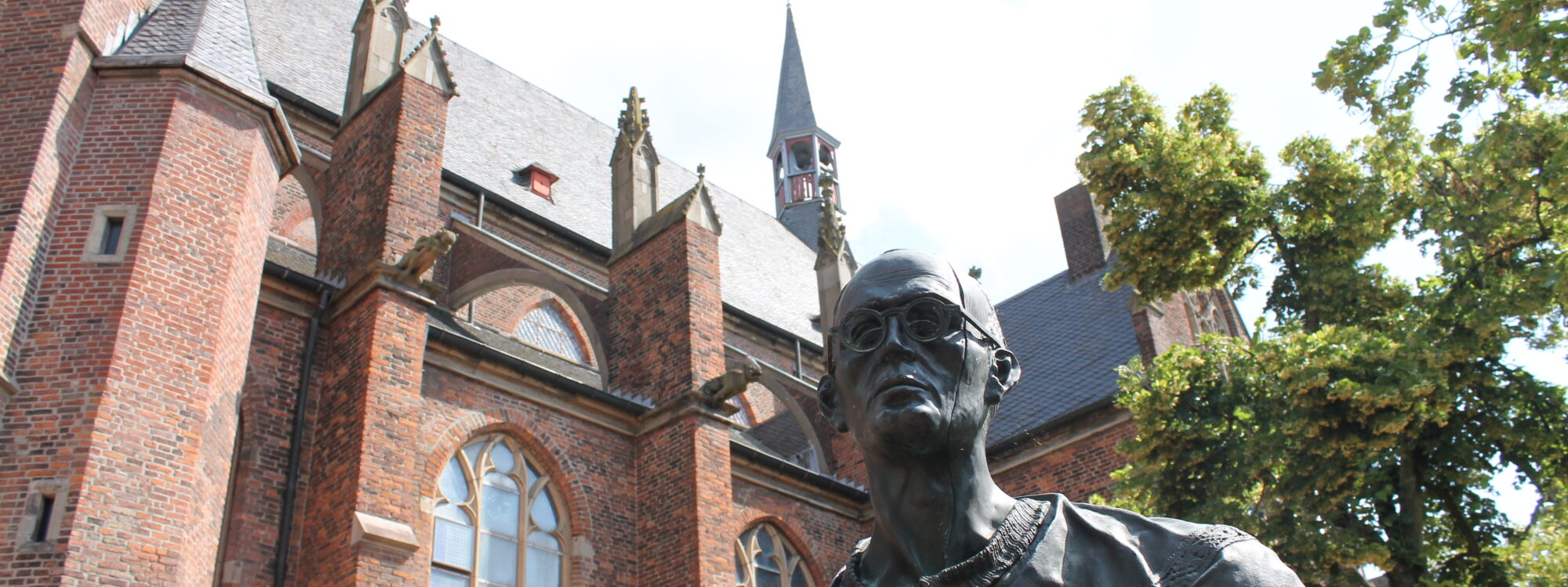 Bronzestatue von Karl Leisner, stehend im Vordergrund. Im Hintergrund die Stiftskirche mit roten Ziegelsteinen und einem hohen Kirchturm.