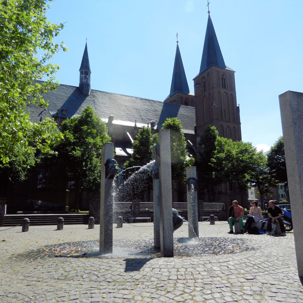 Stiftskirche mit zwei spitzen Türmen im Hintergrund, Narrenbrunnen im Vordergrund mit Wasserfontäne, umgeben von Bäumen und Sitzenden.