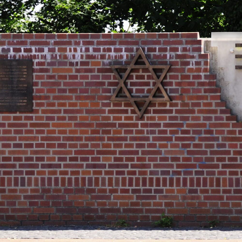 Gedenkstätte am Synagogenplatz mit einem Ziegelmauerstück, einer Davidstern-Skulptur und einer Wand mit Kerzenhaltern.