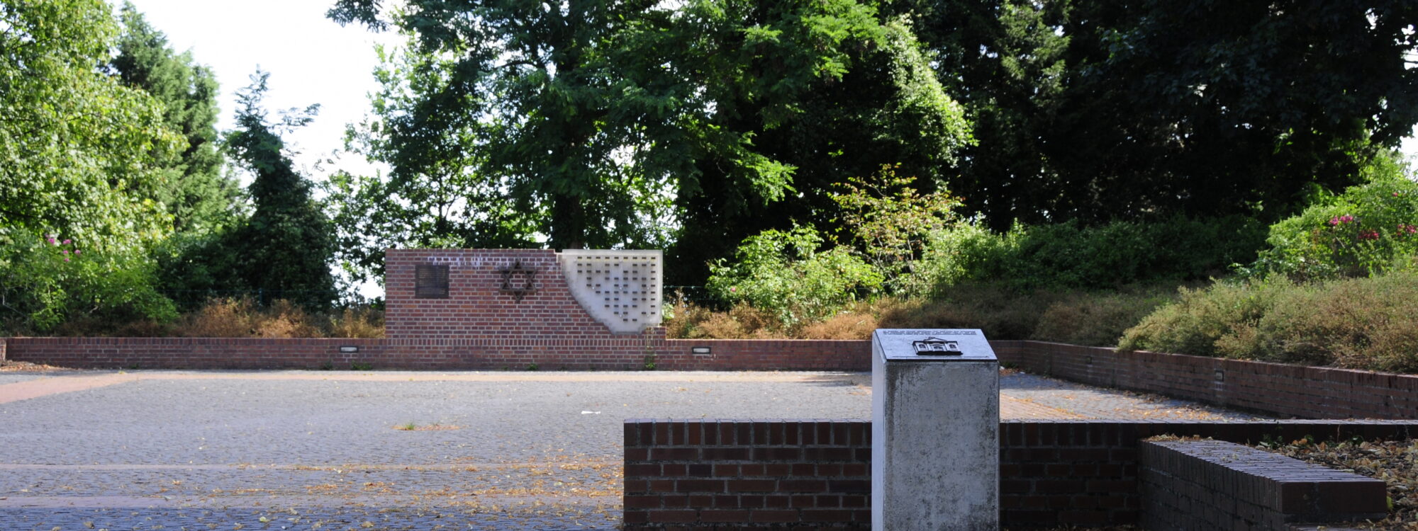 Gedenkstätte am Synagogenplatz mit einem Ziegelmauerstück, einer Davidstern-Skulptur und einer Wand mit Kerzenhaltern.