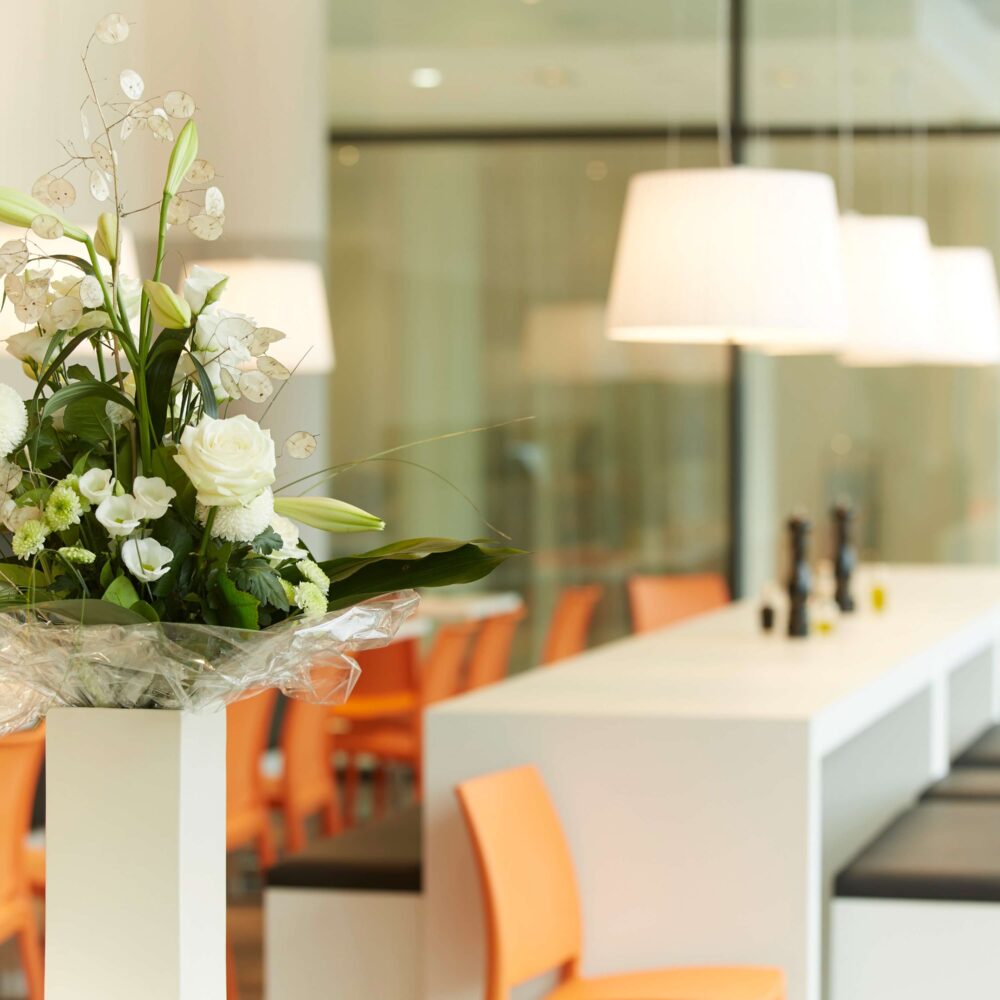 Weiß-grüner Blumenstrauß in Vase vor modernem Raum mit orangen Stühlen und weißen Lampen im elaya hotel kleve