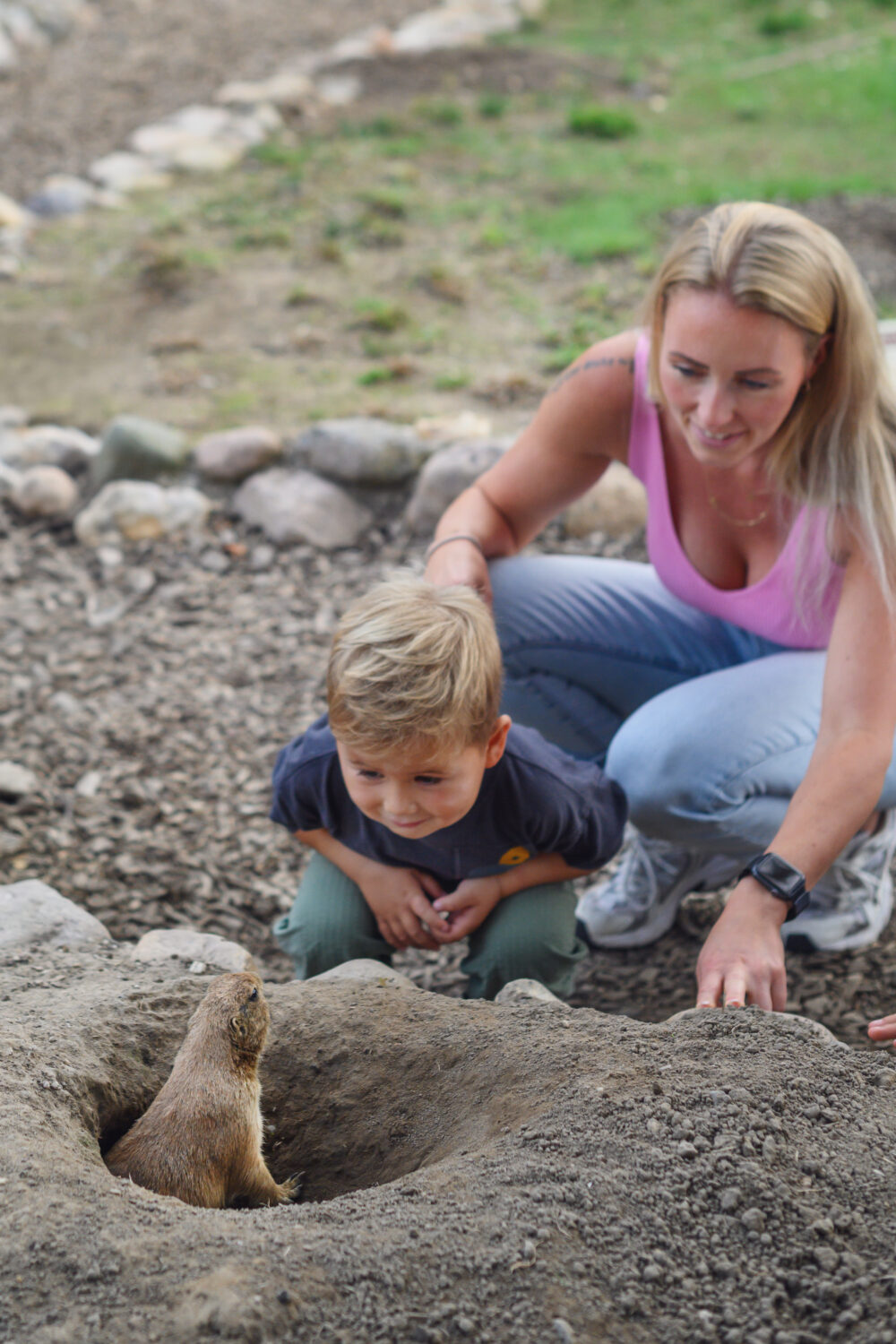 Eine Frau mit langen blonden Haaren und ein Junge mit kurzen blonden Haaren beugen sich über einen Erdhügel. Ein Tier schaut aus einem Loch im Hügel.