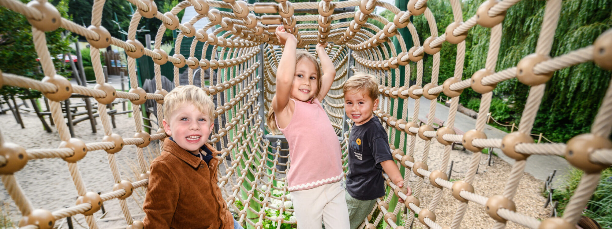 Drei Kinder spielen in einem Klettergerüst aus Seilen. Zwei Kinder sitzen auf der linken und rechten Seite, während ein Kind in der Mitte hängt.