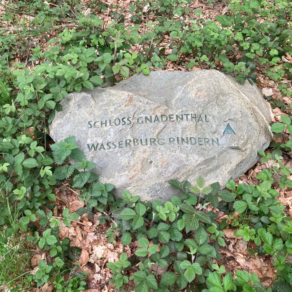 Großer grauer Stein mit den eingravierten Worten 'SCHLOSS GNADENTHAL und WASSERBURG RINDERN' umgeben von grünem Laub und braunen Blättern auf dem Boden.