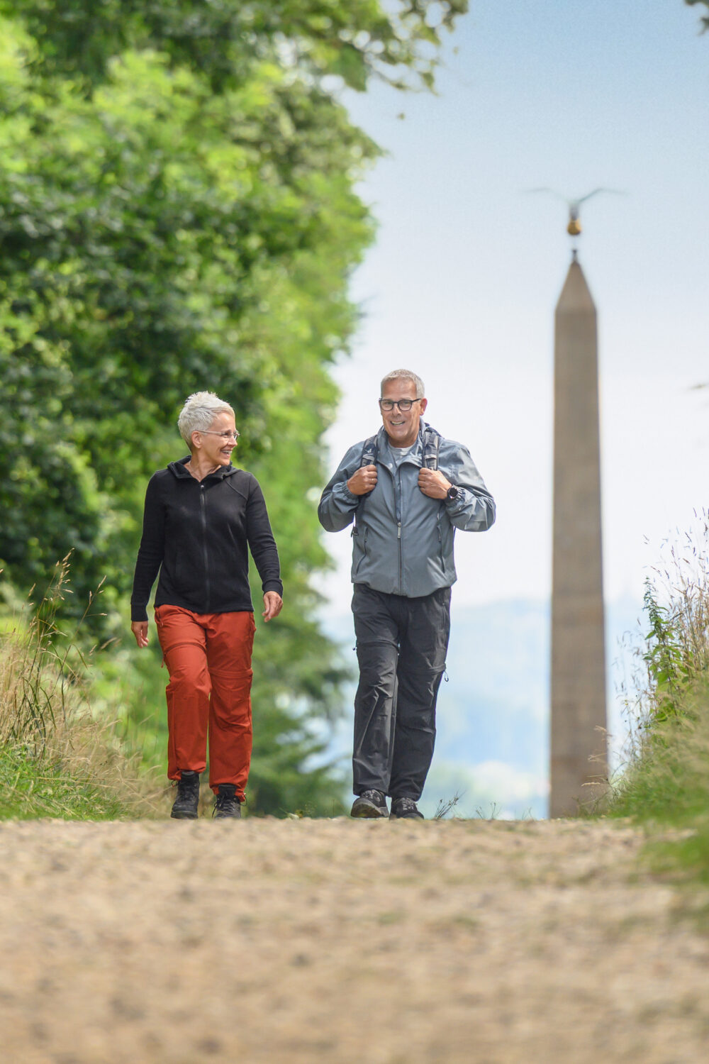 Zwei Personen gehen auf einem Kiesweg durch eine grüne Landschaft. Im Hintergrund steht ein hoher Obelisk.