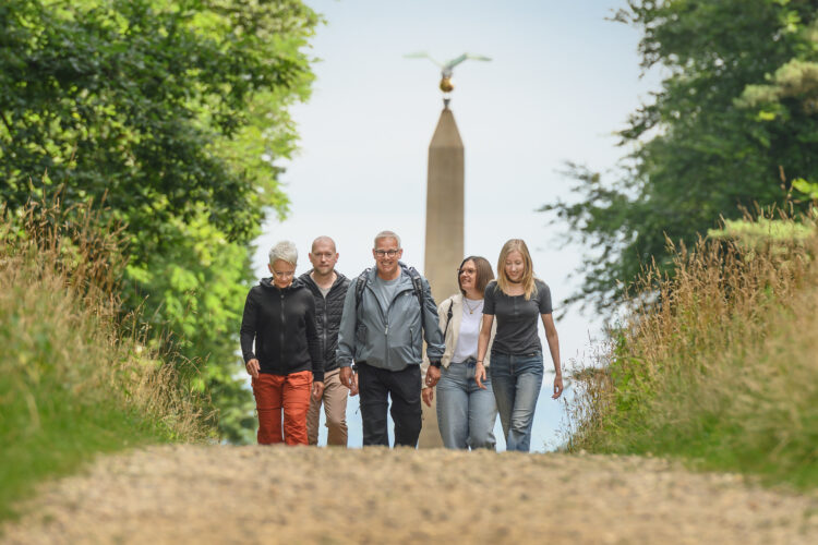 Fünf Personen gehen auf einem Kiesweg, umgeben von Bäumen und hohem Gras. Im Hintergrund steht ein Obelisk mit einer Statue oben.