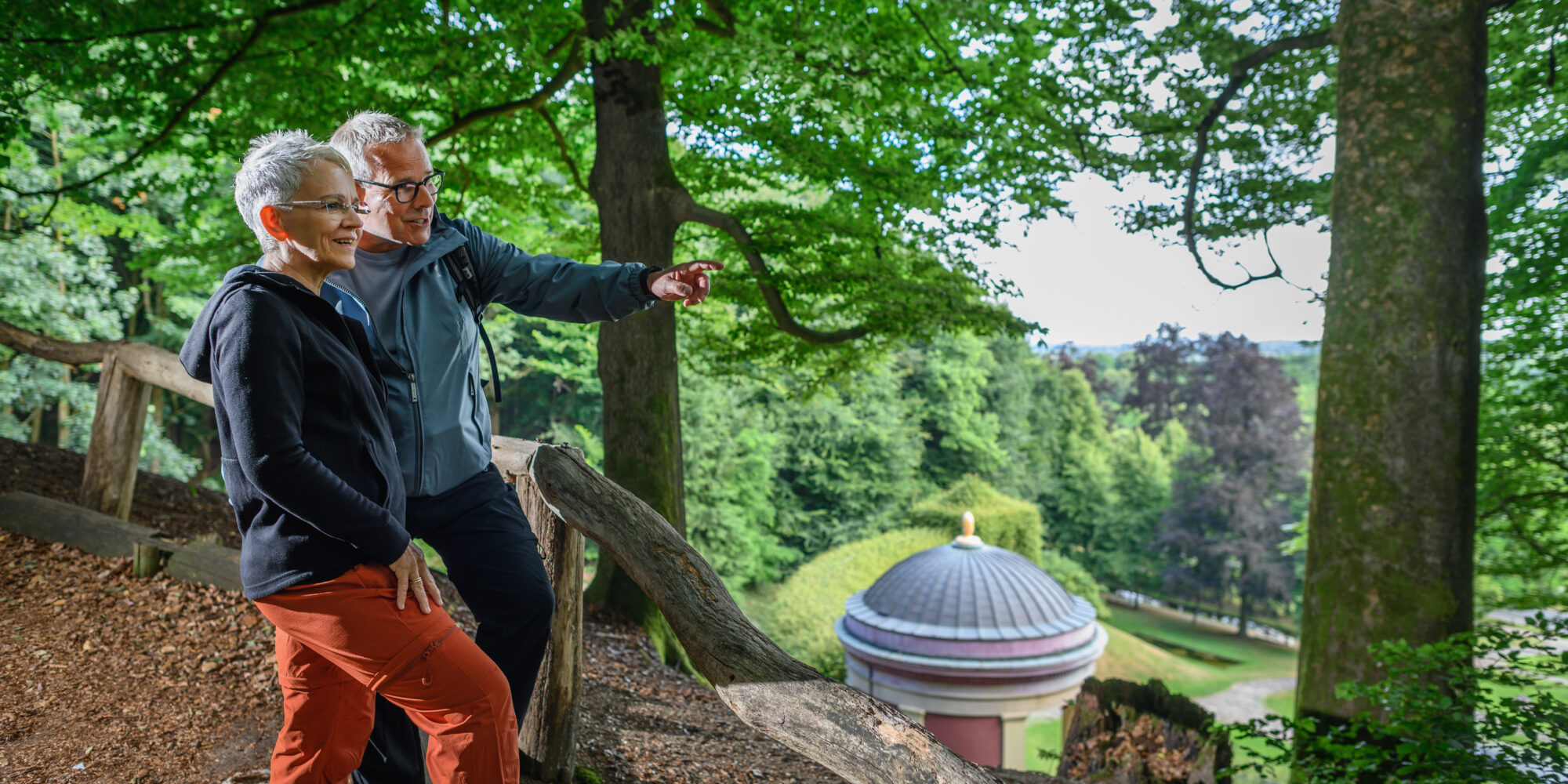 Zwei Personen stehen auf einem Waldweg neben einem Baumstamm, umgeben von grünen Bäumen, mit Blick auf ein rundes Gebäude mit Kuppeldach im Tal.