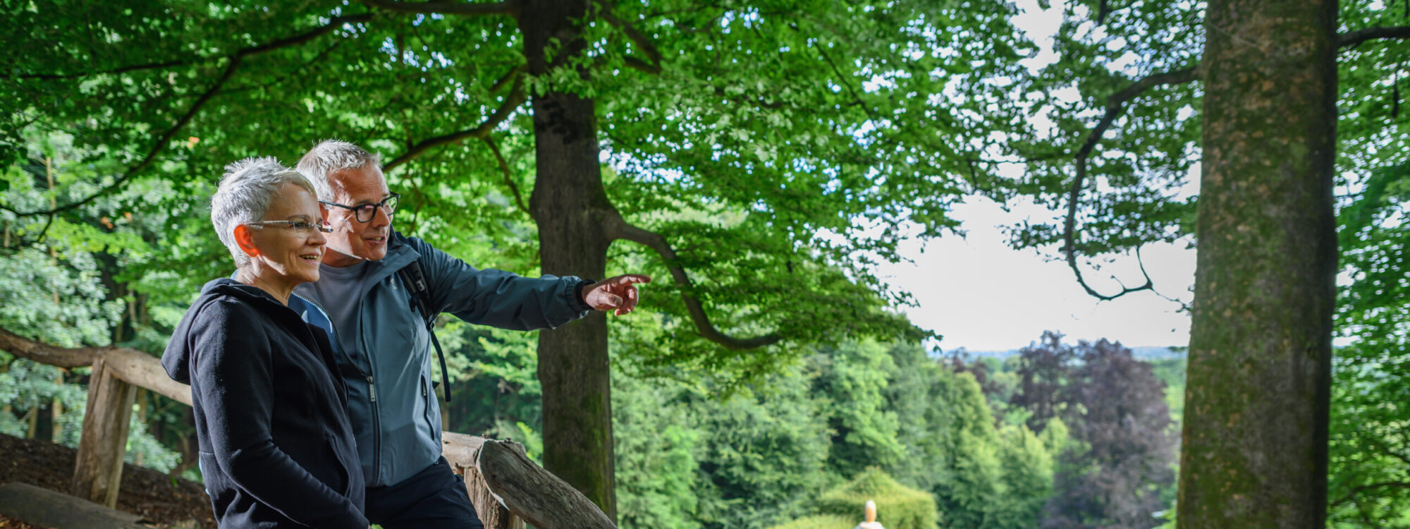 Zwei Personen stehen auf einem Waldweg, eine zeigt mit dem Finger auf etwas im Hintergrund. Im Hintergrund ist ein runder Pavillon mit einer Kuppel sichtbar.