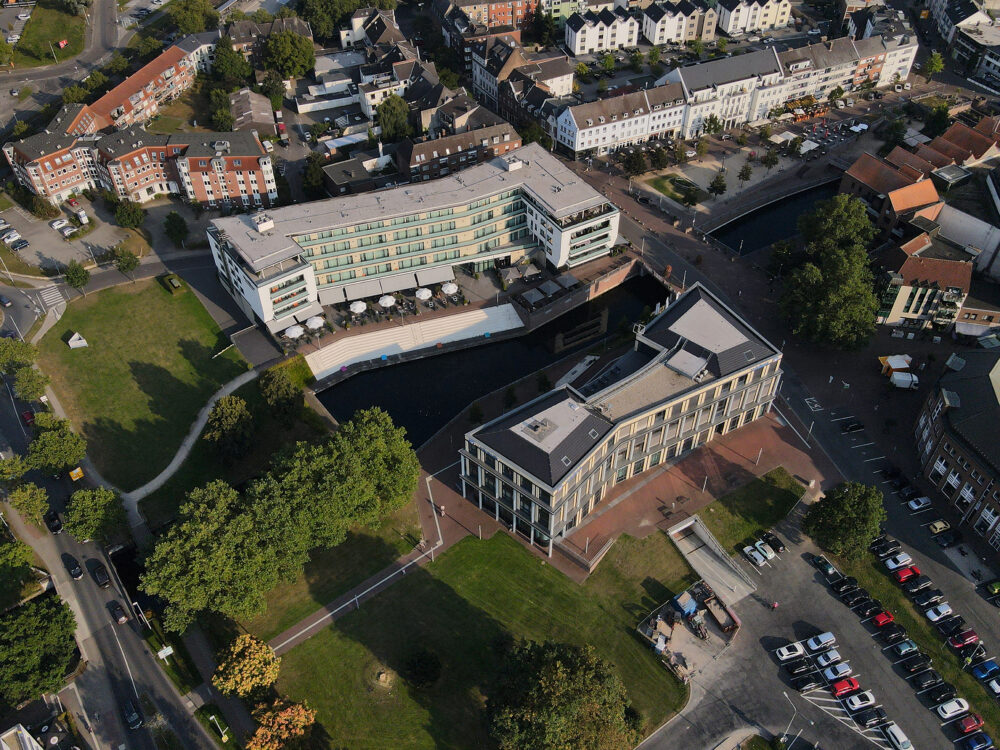 Luftaufnahme des modernen elaya hotel Kleve und der Volksbank mit angrenzendem Park, Parkplatz und umliegender Wohnbebauung
