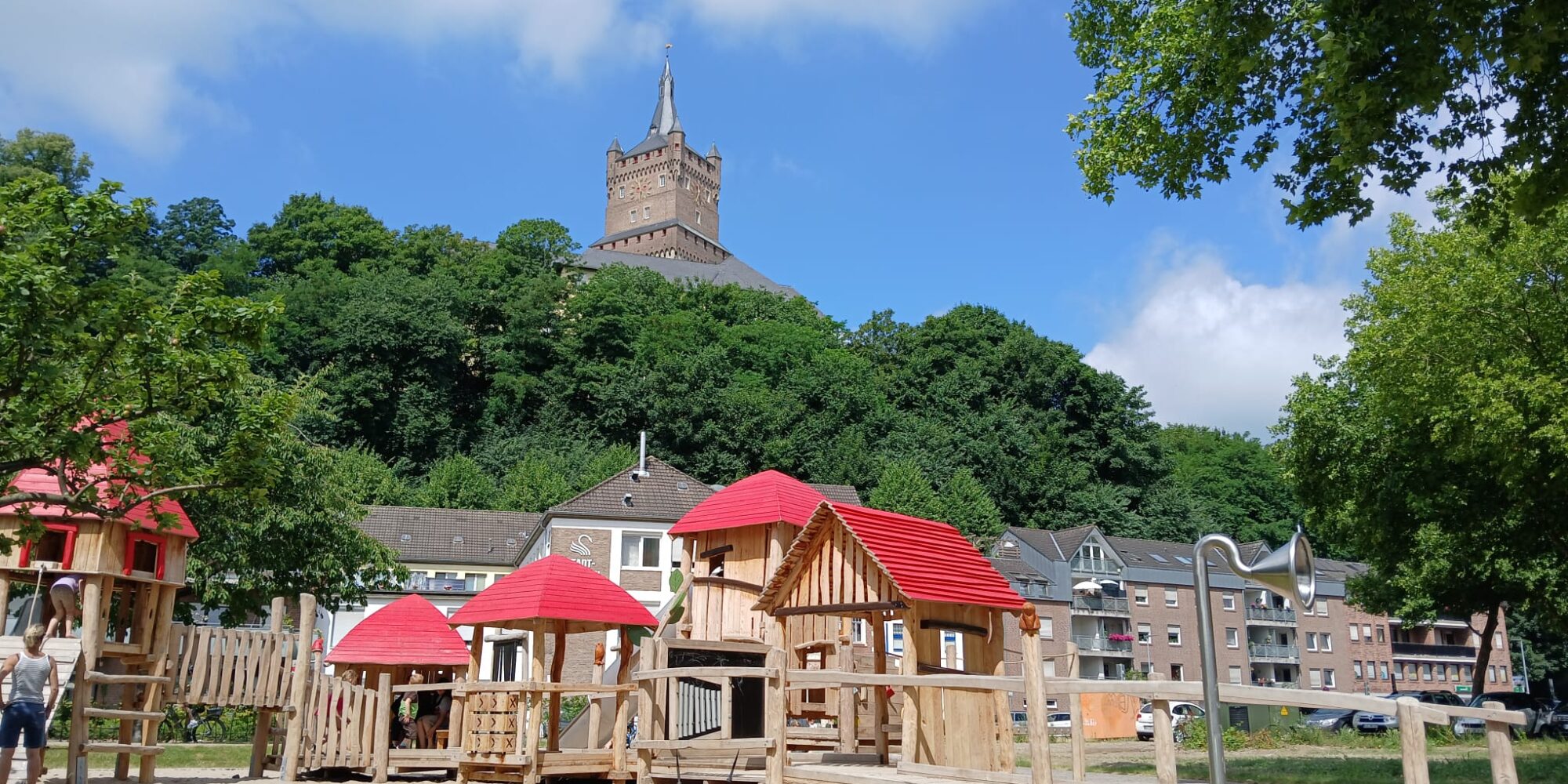 Spielplatz mit mehreren Holzhäusern mit roten Dächern, im Hintergrund eine bewaldete Anhöhe mit einer Burg. Blauer Himmel mit einigen Wolken.