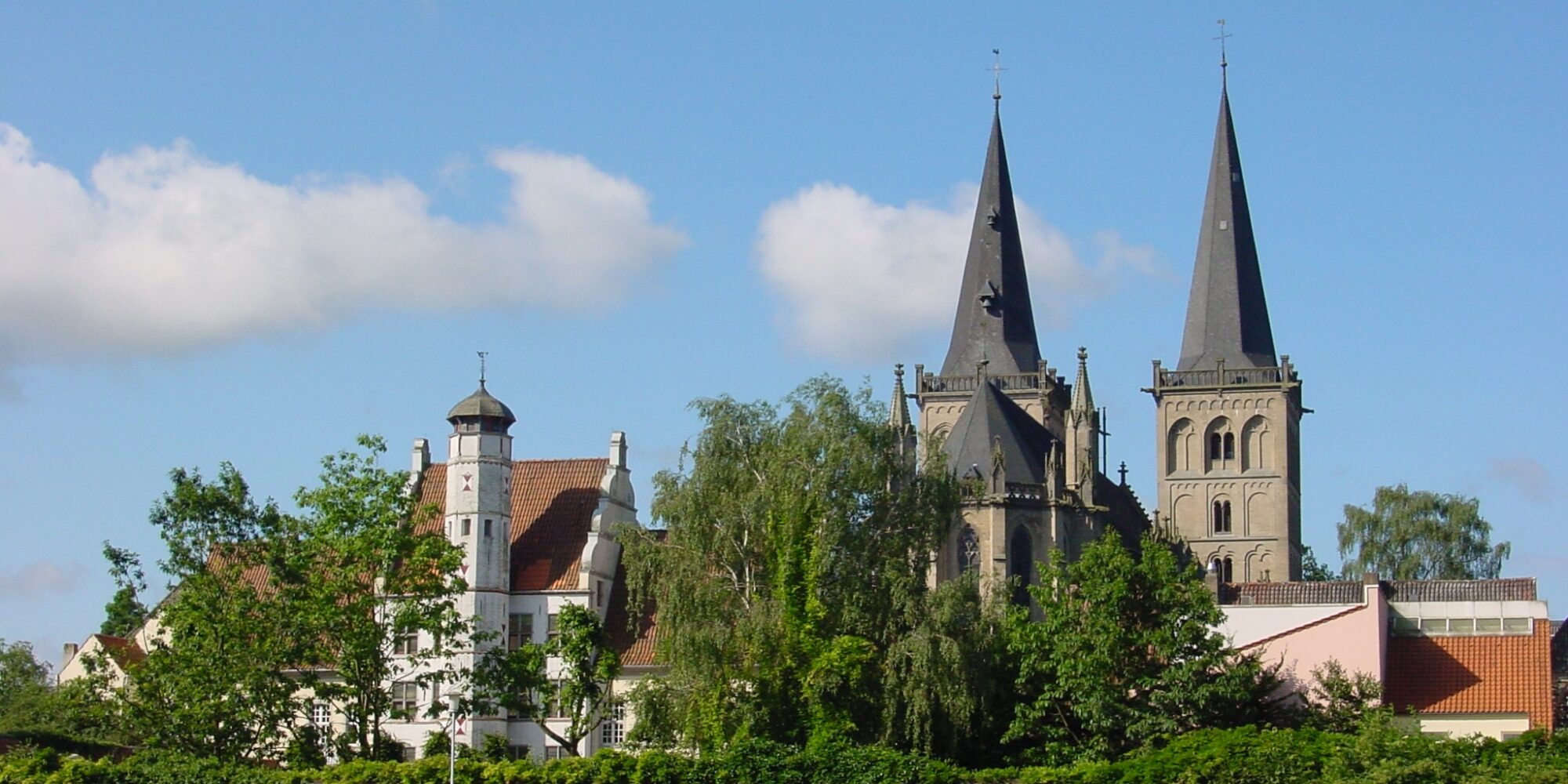 Blick auf die Türme einer gotischen Kirche hinter Bäumen und historischen Gebäuden unter blauem Himmel mit Wolken in Xanten.