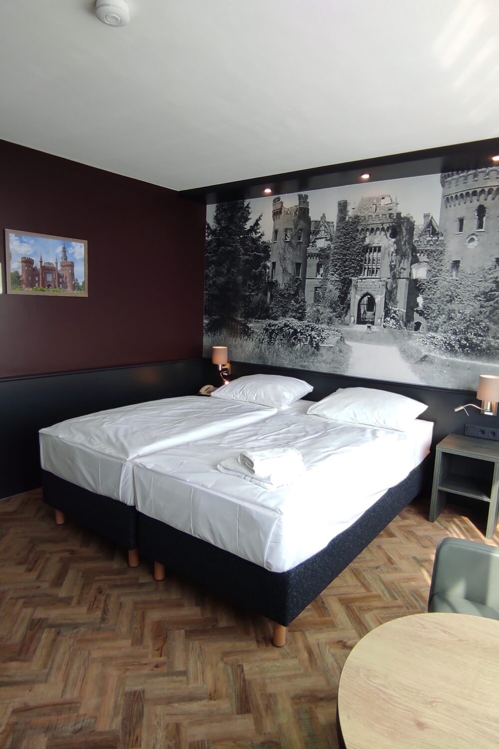 Unterkunft in Kleve. Hotelzimmer mit zwei Einzelbetten, Nachttischen mit Lampen, großem Schwarz-Weiß-Bild einer Burg an der Wand und einem runden Tisch mit zwei Stühlen