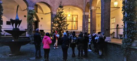 Gruppe von Menschen steht in einem Innenhof mit Weihnachtsbaum und Laternenbeleuchtung, Gästeführerin Birgit van den Boom hält ein Buch und spricht zur Gruppe