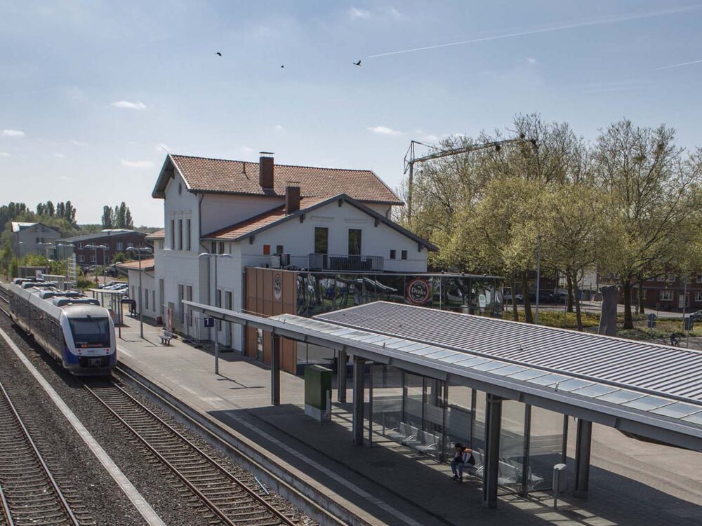 Zug fährt in Klever Bahnhof mit Bahnsteig und wartenden Personen, umgeben von Bäumen und Gebäuden bei klarem Himmel.