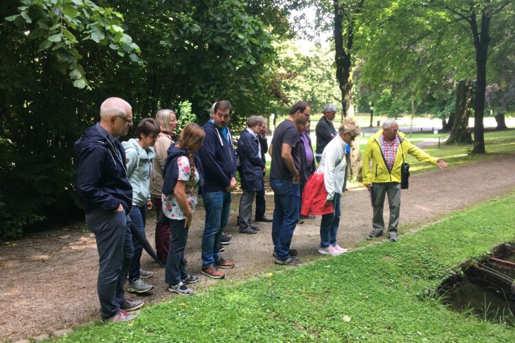 Gruppe von Menschen steht auf einem Weg in den historischen Gartenanlagen von Kleve, ein Mann zeigt auf etwas im Gras neben dem Weg, umgeben von Bäumen.