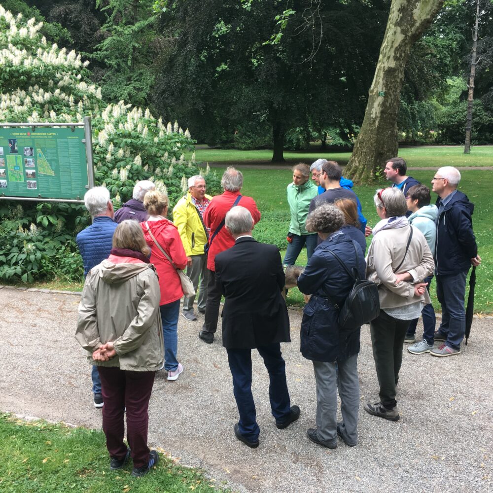 Gruppe von Menschen steht auf einem Weg im Forstgarten vor einer großen Informationstafel mit Text und Bildern, umgeben von Bäumen und Sträuchern.
