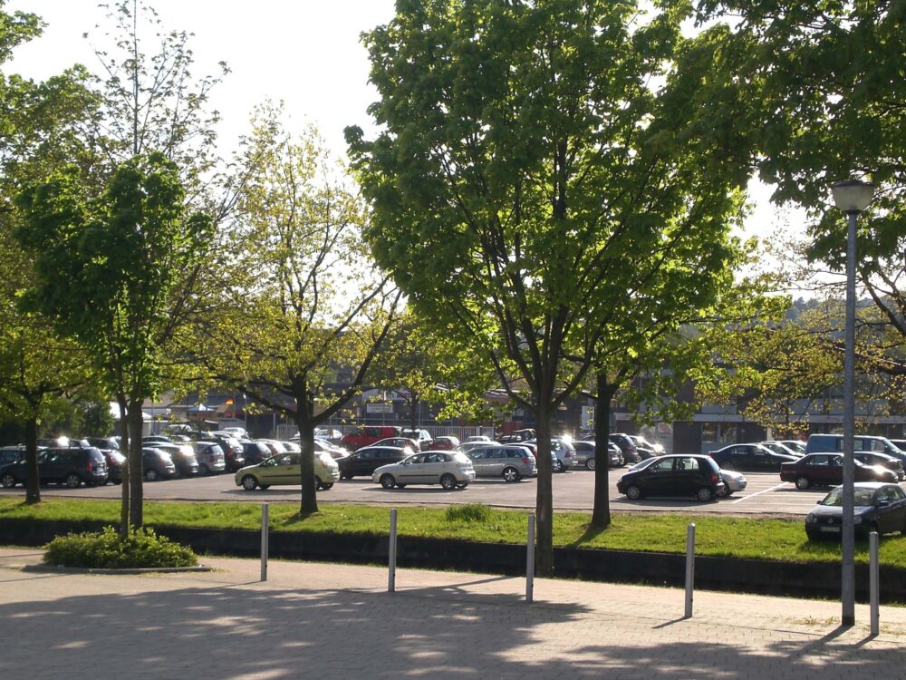 Parkplatz an der Ludwig-Jahn-Straße in Kleve mit mehreren geparkten Autos, Bäume mit grünem Laub und Laterne im Vordergrund bei Tageslicht.