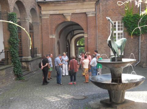 Gruppe von Menschen steht in einem Innenhof mit Kopfsteinpflaster neben einem Brunnen mit Schwan-Skulptur aus Metall.