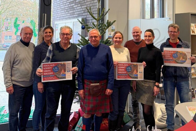 Gruppe von acht Personen steht vor einem Weihnachtsbaum, zwei halten Plakate mit dem Text 'NIKOLAUS KOMMT ÜBERS WASSER'.