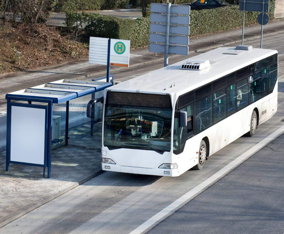 Weißer Linienbus steht an einer Haltestelle mit blauem Wartehäuschen und Haltestellenschild.