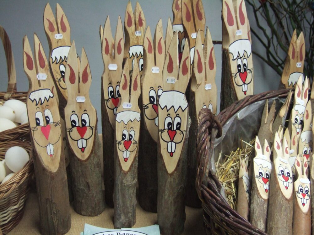 Mehrere aus Holzstämmen gefertigte Osterhasen mit bemalten Gesichtern und roten Nasen, stehend in einem Korb und auf einer Fläche.