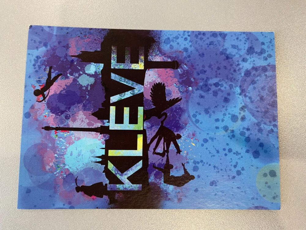 Postkarte mit dem Schriftzug 'KLEVE' in Gelb vor schwarzer Silhouette von Gebäuden und Figuren auf blau-lila Hintergrund mit Farbspritzern.