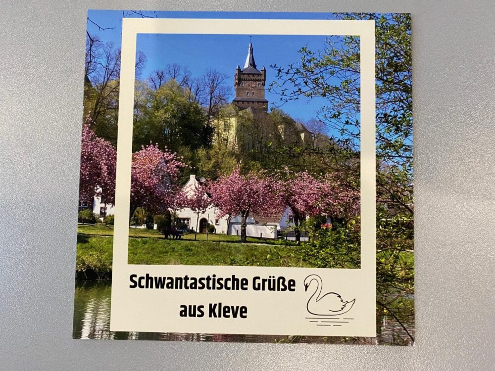 Postkarte mit Kirchturm hinter blühenden Kirschbäumen, darunter Text 'Schwantastische Grüße aus Kleve' und eine Schwan-Illustration.
