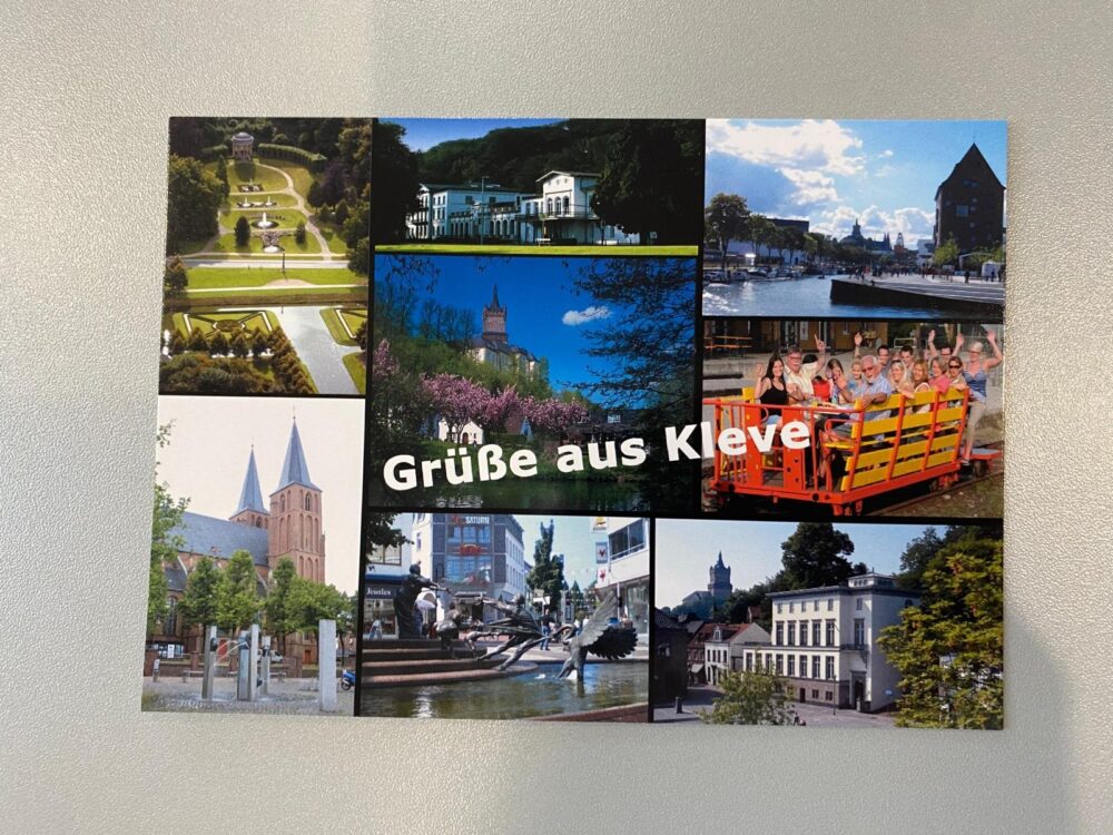 Postkarte mit mehreren Fotos von Kleve: Parkanlage, weiße Villa, Fluss mit Boot, Kirche, Brunnen mit Skulpturen, Stadtansichten und Menschen auf einem gelben Ausflugsboot. Text 'Grüße aus Kleve' in weißer Schrift.