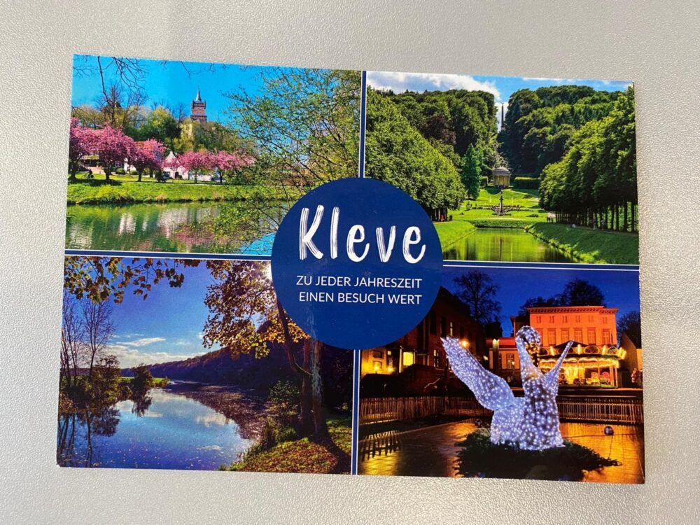 Postkarte mit vier Fotos von Kleve: blühende Bäume am Wasser, grüne Allee mit Pavillon, Flusslandschaft im Herbst, beleuchtete Schwanenskulptur am Abend. Text in blauem Kreis: 'Kleve zu jeder Jahreszeit einen Besuch wert'.