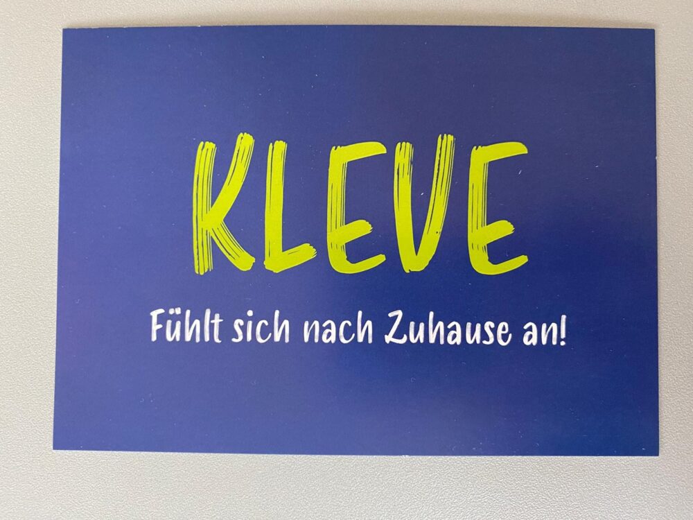 Postkarte mit dunkelblauem Hintergrund und grünem Schriftzug 'KLEVE', darunter weißer Text 'Fühlt sich nach Zuhause an!'
