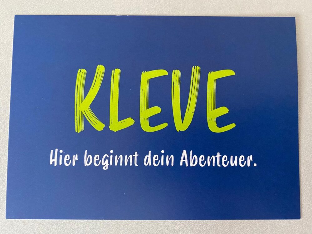 Postkarte mit blauem Hintergrund und grünem Schriftzug 'KLEVE', darunter weißer Text 'Hier beginnt dein Abenteuer.'