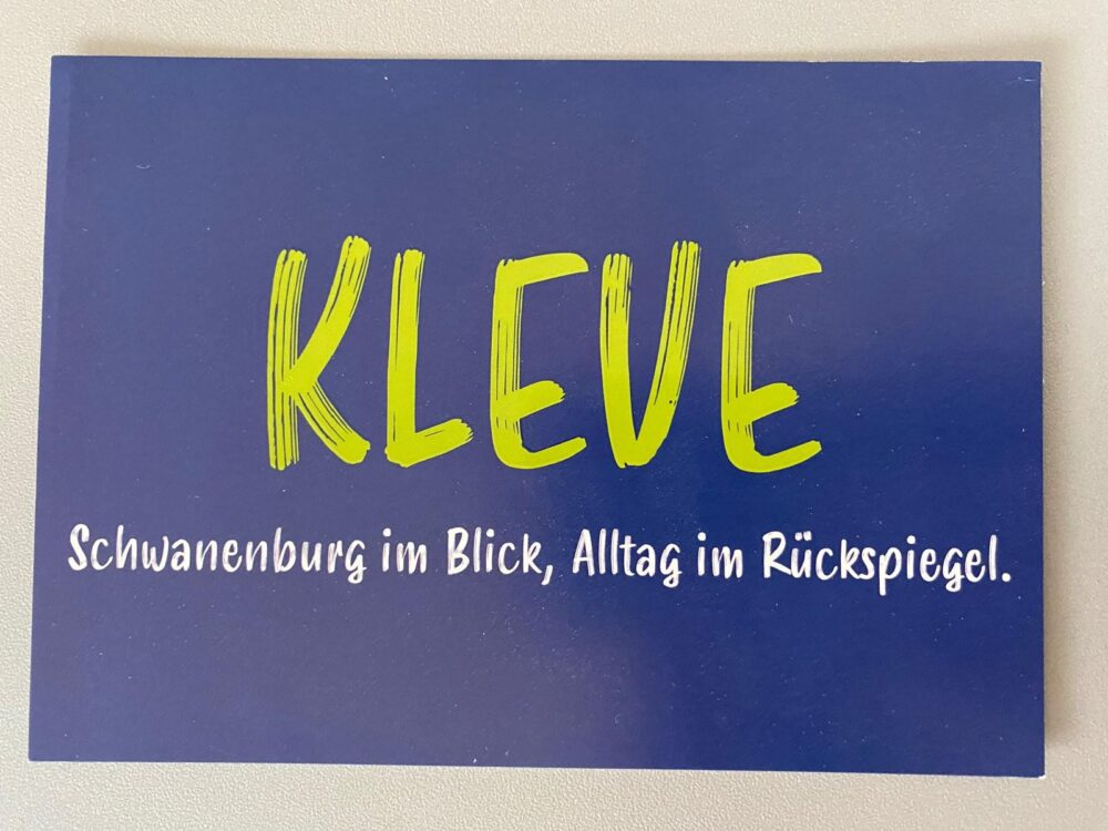 Blaues Postkartenmotiv mit grünem Schriftzug 'KLEVE' und weißer Schrift darunter: 'Schwanenburg im Blick, Alltag im Rückspiegel.'