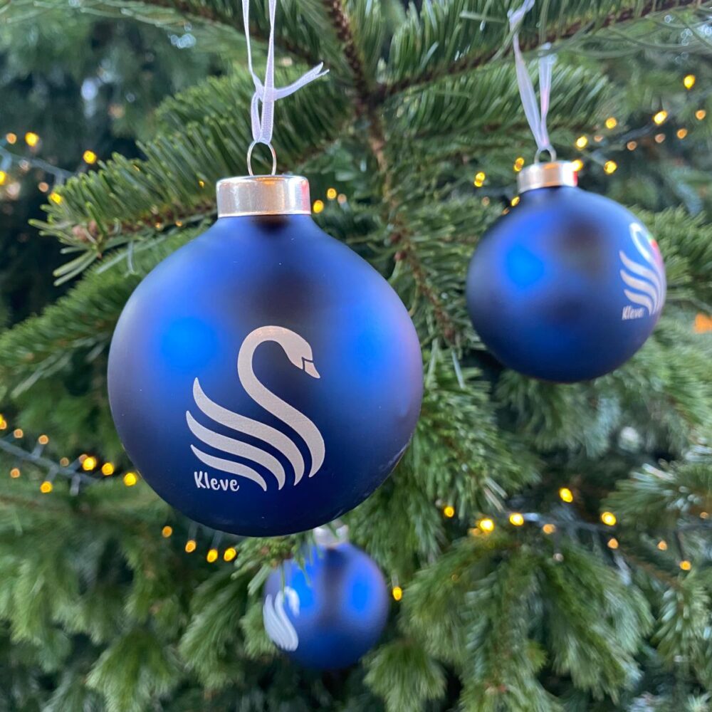 Drei blaue Weihnachtskugeln mit weißem Schwanenmotiv und Schriftzug 'Kleve' hängen an einem Tannenzweig, im Hintergrund Lichterkette.