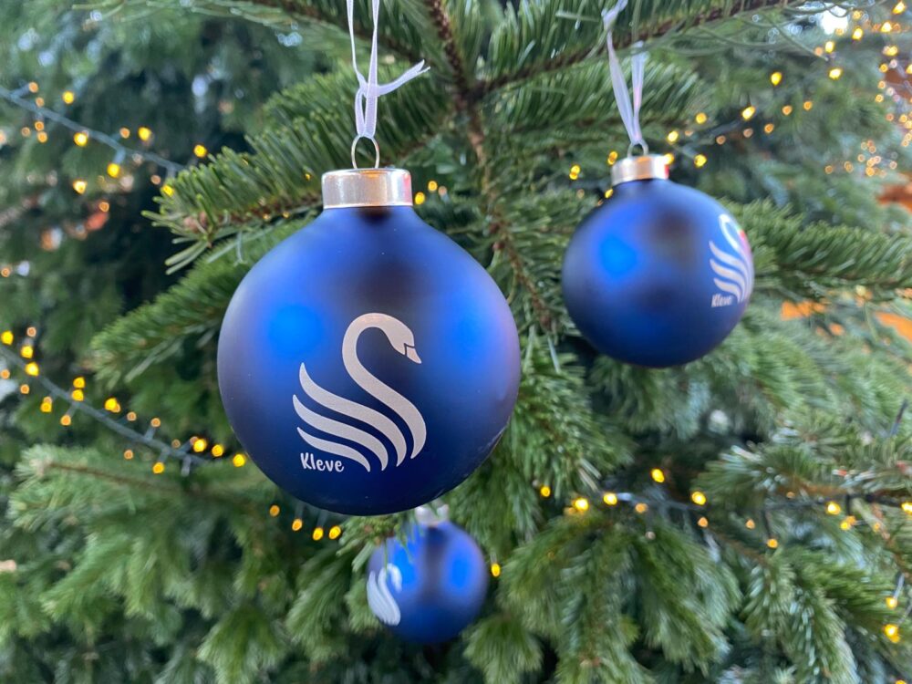 Drei blaue Weihnachtskugeln mit weißem Schwanenmotiv und Schriftzug 'Kleve' hängen an einem Tannenzweig, im Hintergrund Lichterkette.