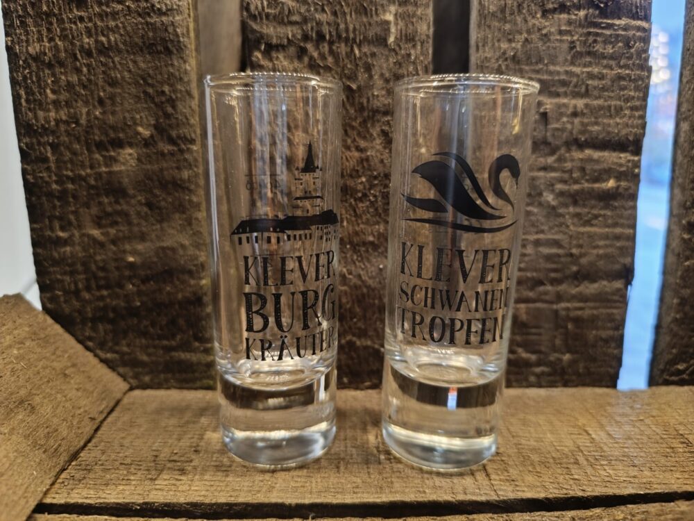 Zwei klare Schnapsgläser mit schwarzem Aufdruck, links mit Kirchturm und Text 'KLEVER BURG KRÄUTER', rechts mit Schwan und Text 'KLEVER SCHWANEN TROPFEN'.