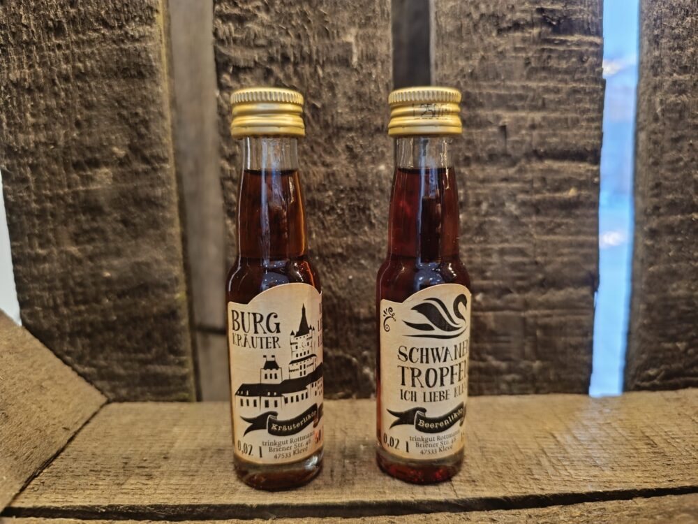 Zwei kleine braune Flaschen mit goldfarbenen Verschlüssen, Etiketten mit schwarzer Schrift und Illustrationen, stehen auf Holz vor dunklem Hintergrund.