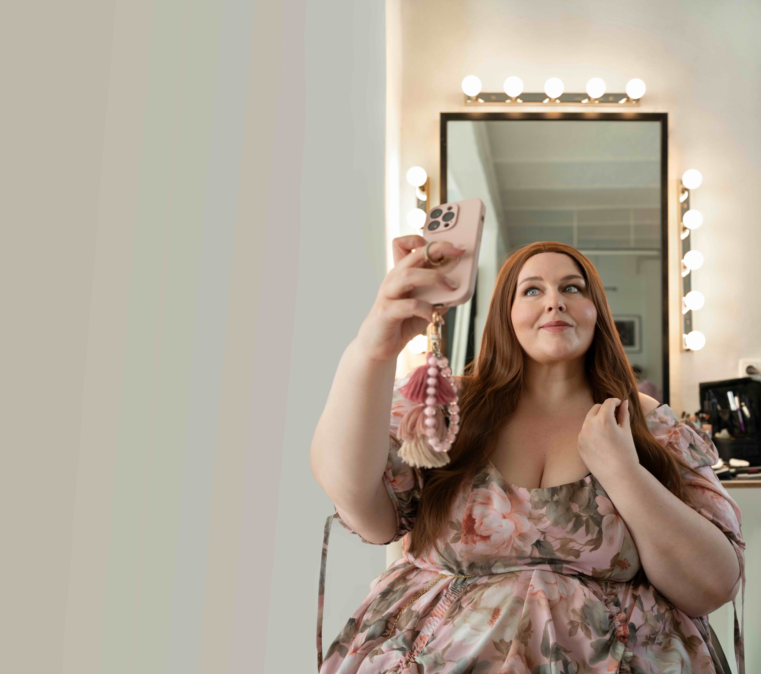 Nicole Jäger in einem geblümtem Kleid macht ein Selfie vor beleuchtetem Spiegel und hält ein Smartphone mit rosa Hülle und Perlenanhänger hoch.