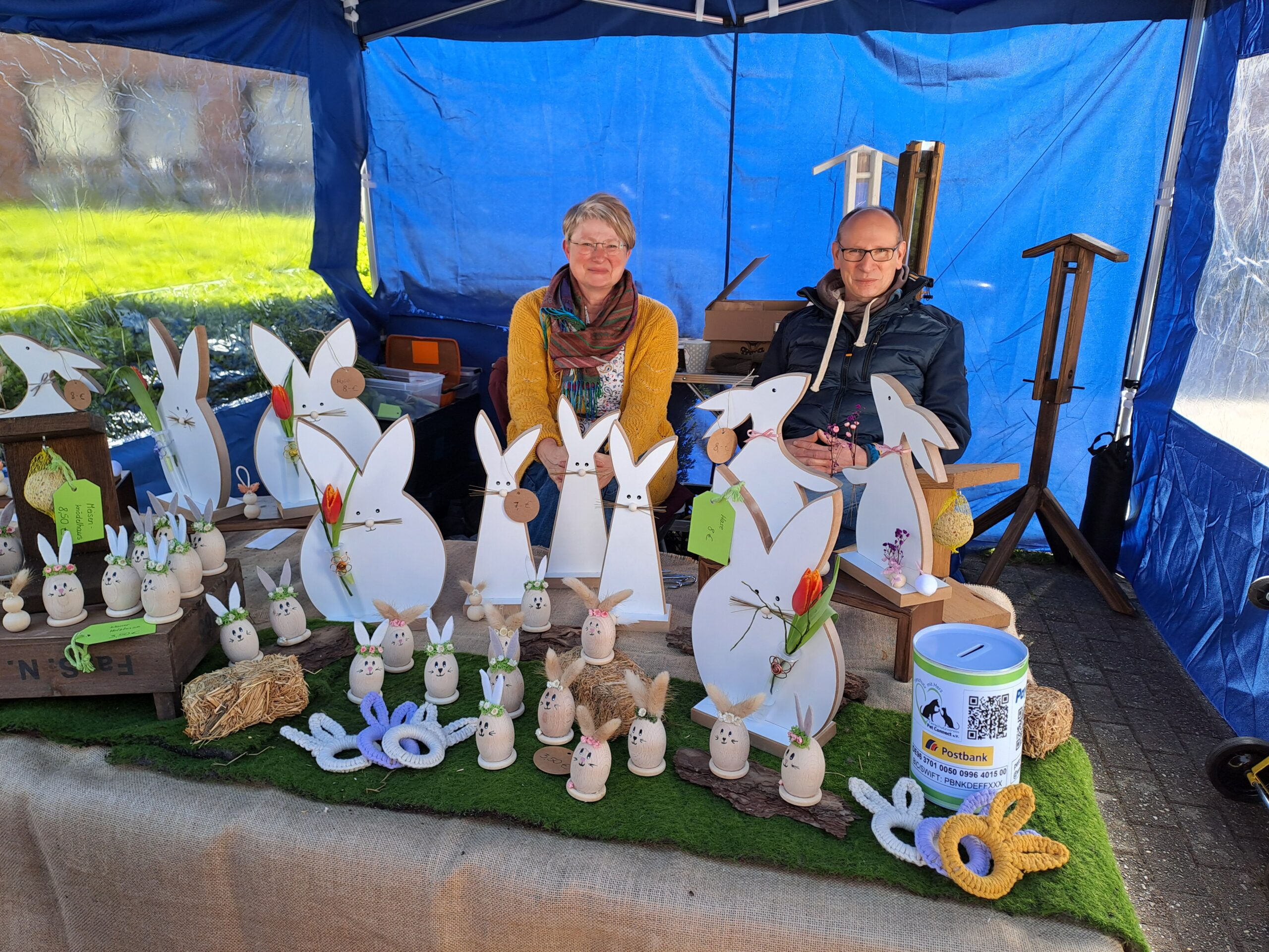 Zwei Personen sitzen hinter einem Stand mit verschiedenen weißen Osterhasenfiguren aus Holz und Keramik, dekoriert mit Karotten und Blumen.