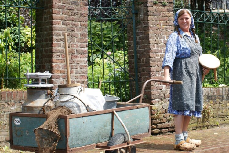 Frau in traditioneller Kleidung mit Holzschuhen steht neben einem mit Milchkannen und Netzen beladenen Handwagen vor einem Backsteinzaun.