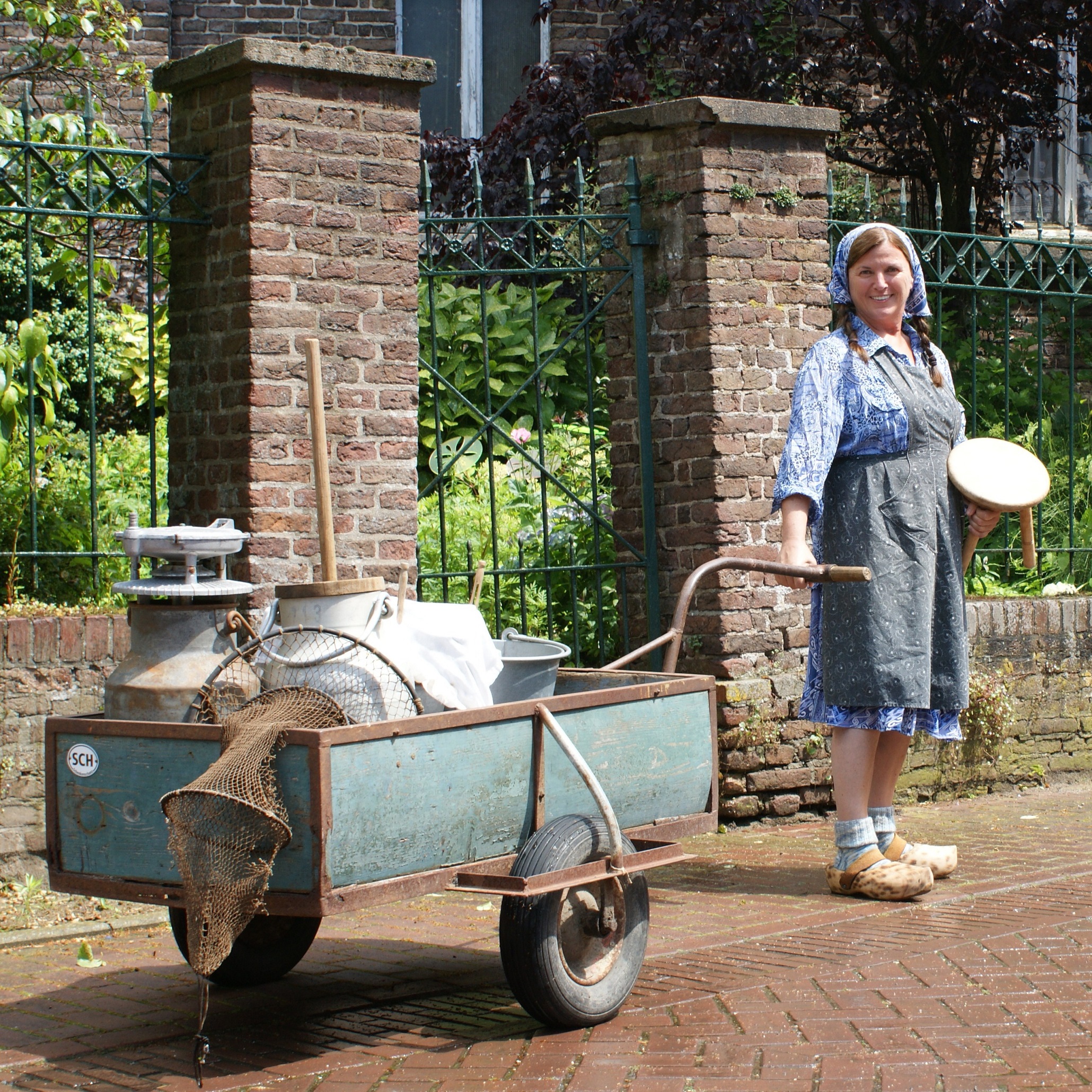 Frau in traditioneller Kleidung mit Holzschuhen steht neben einem mit Milchkannen und Netzen beladenen Handwagen vor einem Backsteinzaun.