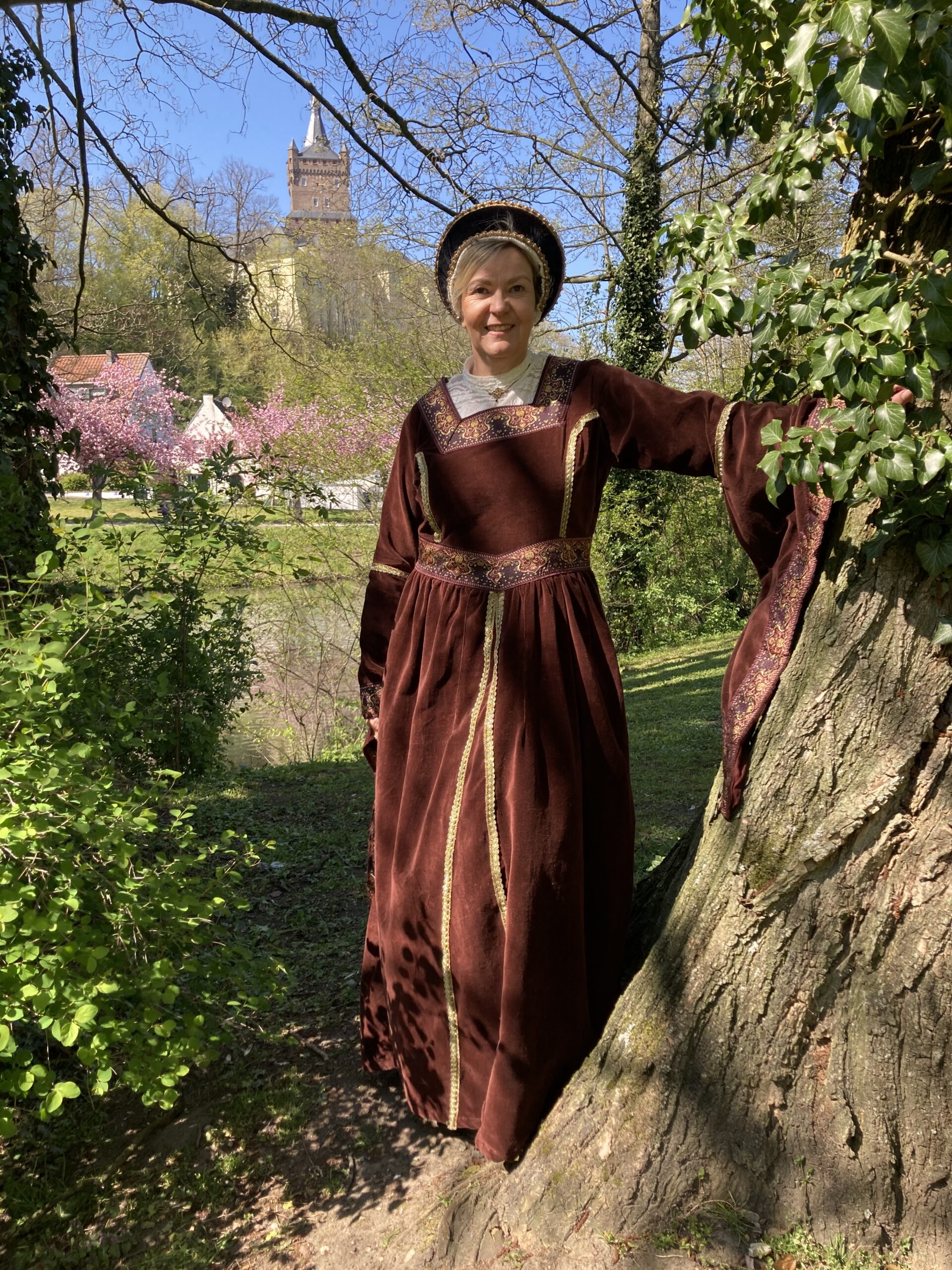 Stadtführerin Birgit van den Boom in historischer, langärmliger Robe mit goldenen Verzierungen lehnt an einem Baum in einem Park mit Schloss im Hintergrund.