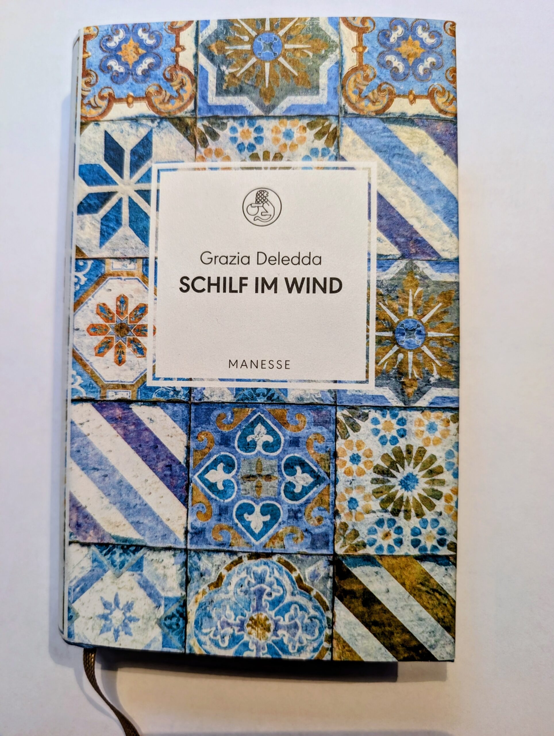 Buchdeckel mit Muster aus blauen, weißen und goldenen Fliesen, mittig weißes Rechteck mit Text 'Grazia Deledda SCHILF IM WIND MANESSE'.