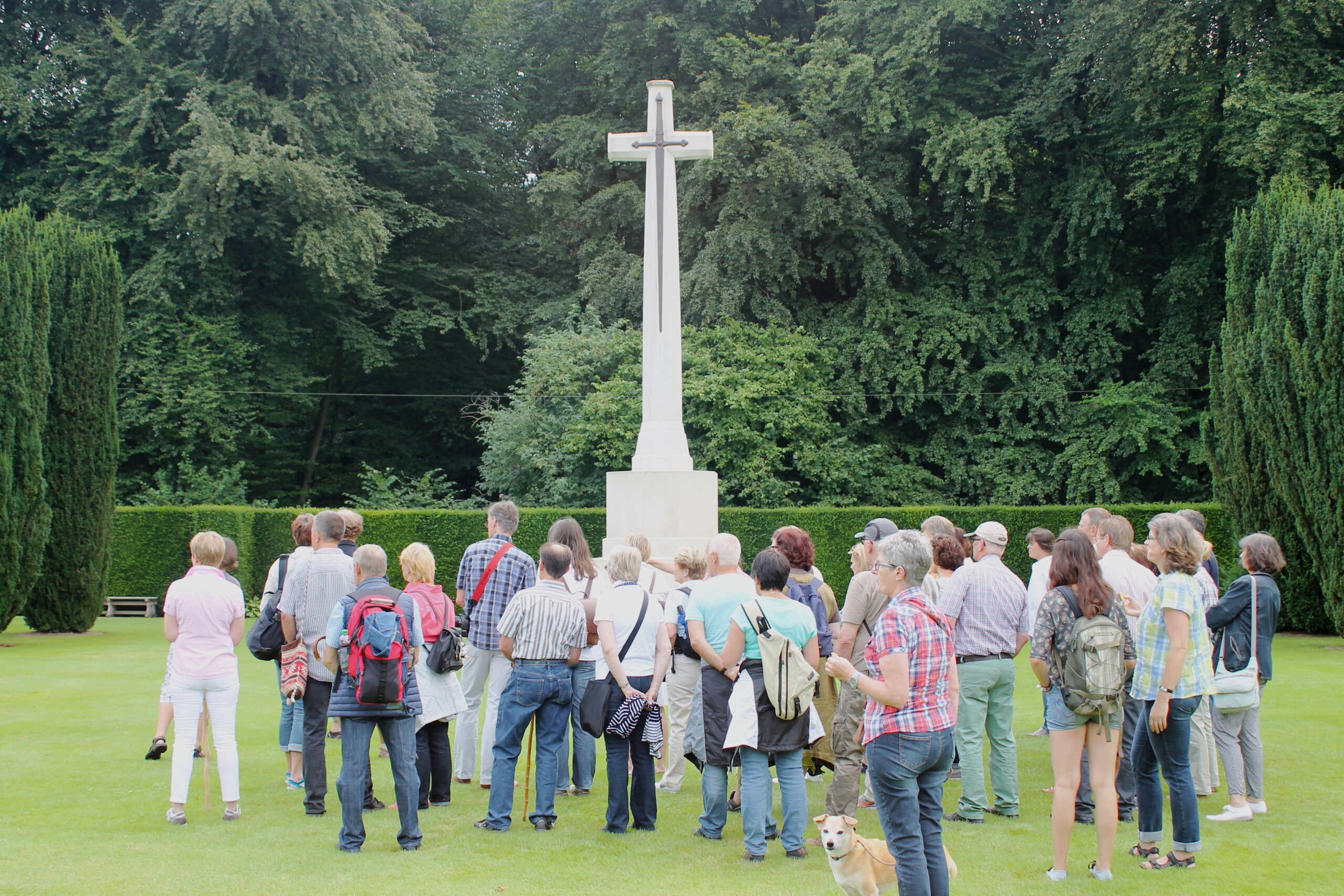 Gruppe von Menschen steht auf einer Rasenfläche vor einem hohen Kreuz auf einem Sockel, umgeben von Bäumen und Hecken