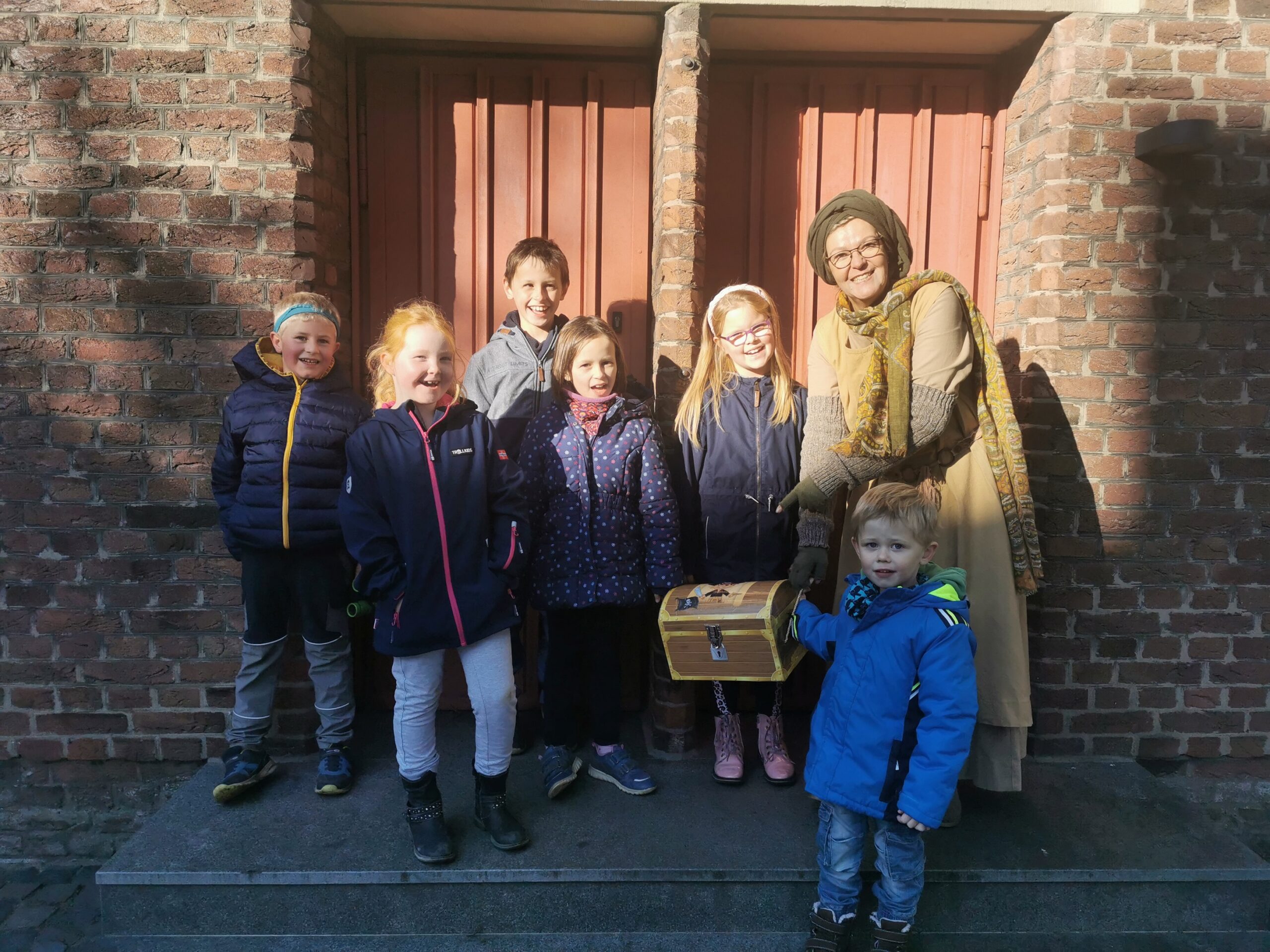 Eine Gruppe von Kindern und Gästeführerin Birgit van den Boom stehen vor einer Holztür aus Backsteinmauerwerk und halten eine Schatztruhe