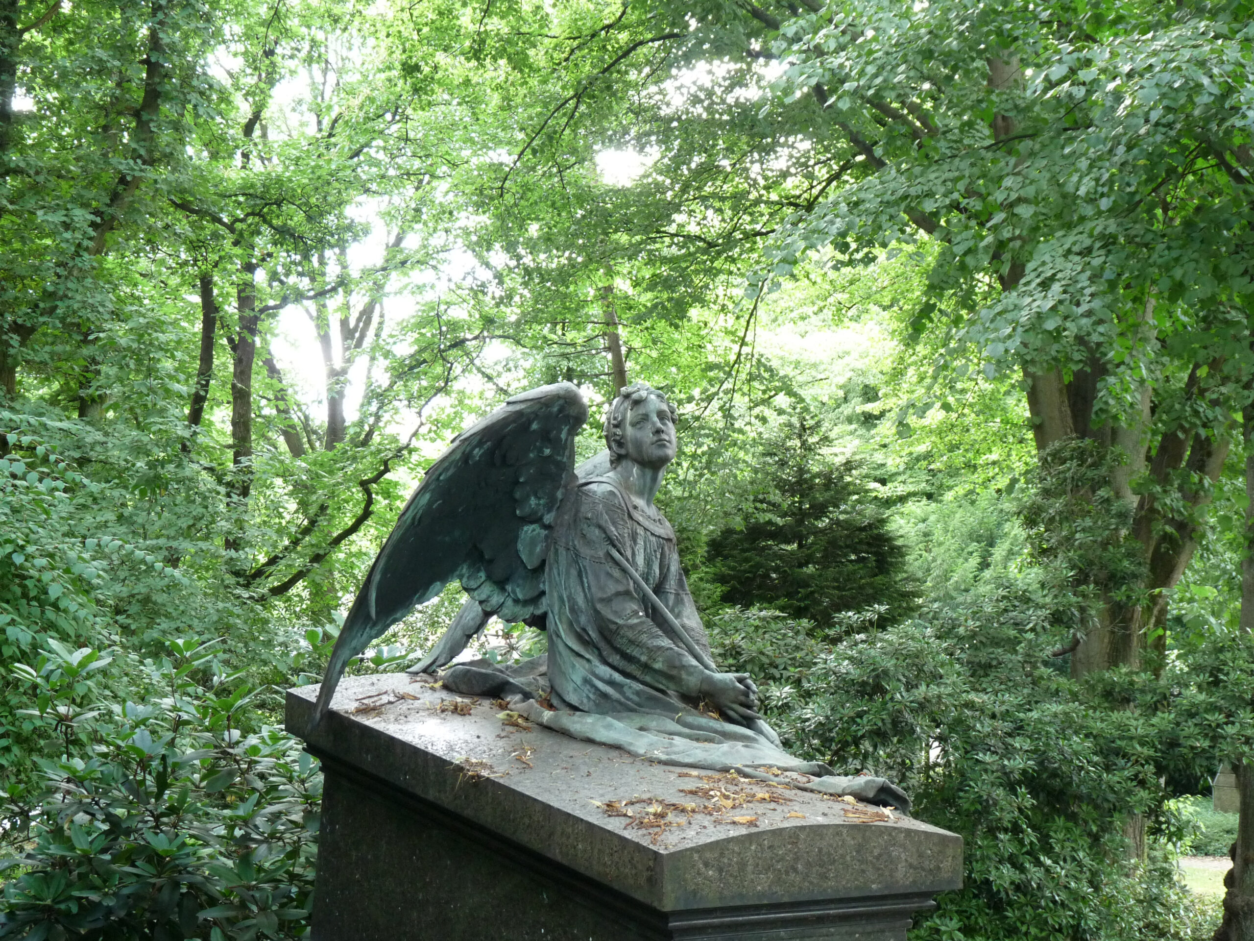 Bronzefigur eines Engels mit ausgebreiteten Flügeln, der auf einem schrägen Steinsockel sitzt, umgeben von Bäumen und Grün.